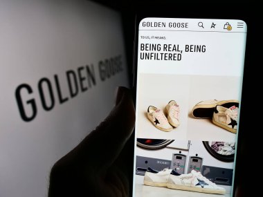 Stuttgart, Almanya - 03-05-2024: İtalyan spor ayakkabı şirketi Golden Goose S.P.A. 'nın internet sayfası ile logo önünde cep telefonu tutan kişi. Telefon ekranının merkezine odaklan.