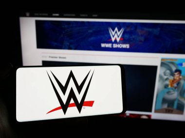 Stuttgart, Germany - 03-07-2024: Person holding mobile phone with logo of promotion company World Wrestling Entertainment (WWE) in front of web page. Focus on phone display. clipart