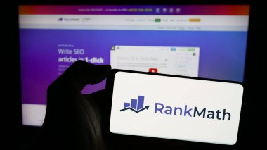 Stuttgart, Almanya - 03-12-2024: Indian WordPress SEO şirketi Rank Math 'in logosuna sahip cep telefonu sahibi kişi iş sayfasının önünde. Telefon ekranına odaklan.