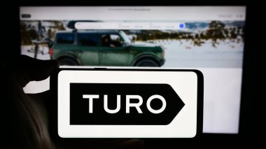 Stuttgart, Almanya - 03-18-2024: İş dünyasının web sayfası önünde ABD 'li yaşıtlar arası otomobil paylaşım şirketi Turo Inc.' nin logosuyla cep telefonu bulunduran kişi. Telefon ekranına odaklan.