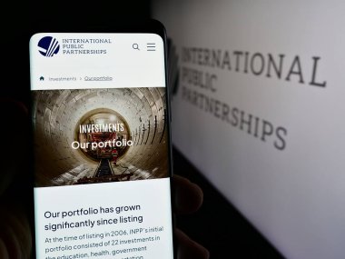 Stuttgart, Almanya - 03-20-2024: İngiliz şirketi International Public Partnerships Limited 'in logolu web sayfası ile cep telefonu tutan kişi. Telefon ekranının merkezine odaklan.