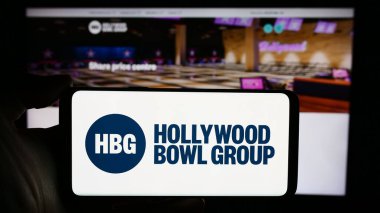 Stuttgart, Germany - 03-20-2024: Person holding cellphone with logo of British bowling company Hollywood Bowl Group plc in front of business webpage. Focus on phone display. clipart