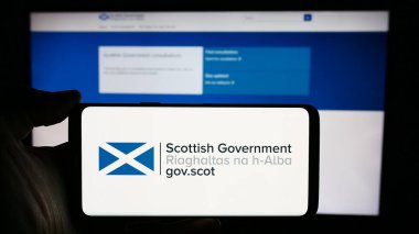 Stuttgart, Germany - 03-25-2024: Person holding mobile phone with logo of Scottish Government (Riaghaltas na h-Alba) in front of web page. Focus on phone display. clipart