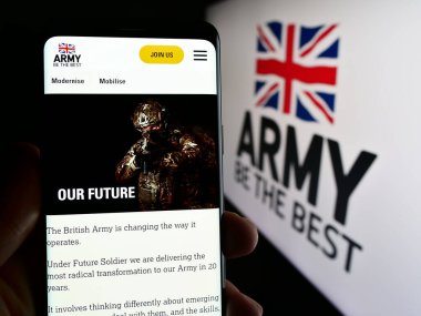 Stuttgart, Germany - 03-25-2024: Person holding mobile phone with web page of United Kingdom ground force British Army in front of logo. Focus on center of phone display. clipart