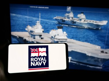 Stuttgart, Germany - 03-25-2024: Person holding mobile phone with logo of United Kingdom naval warfare Royal Navy (RN) in front of web page. Focus on phone display. clipart