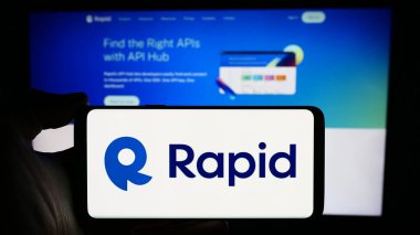 Stuttgart, Almanya - 03-28-2024: Üzerinde US API merkez şirketi R Software Inc. (RapidAPI) 'nin logosu bulunan cep telefonu sahibi kişi iş sayfası önünde. Telefon ekranına odaklan.