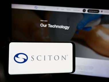 Stuttgart, Germany - 04-05-2024: Person holding smartphone with logo of US medical device company Sciton Inc. in front of website. Focus on phone display. clipart