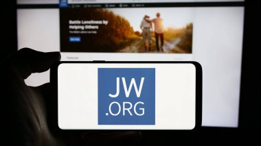 Stuttgart, Germany - 04-15-2024: Person holding cellphone with logo of religious organisation Jehovah's Witnesses in front of webpage. Focus on phone display. clipart