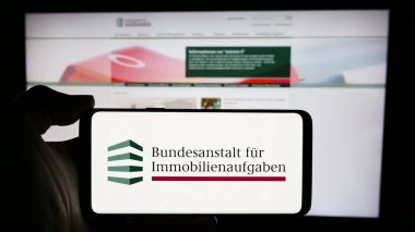 Stuttgart, Germany - 04-15-2024: Person holding cellphone with logo of German agency Bundesanstalt fuer Immobilienaufgaben (BImA) in front of webpage. Focus on phone display. clipart