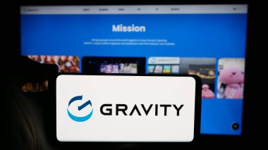 Stuttgart, Almanya - 04-17-2024: Güney Koreli video oyunu şirketi Gravity Co. Ltd. nin logosuyla cep telefonu tutan kişi iş sayfasının önünde. Telefon ekranına odaklan.