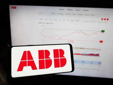 Stuttgart, Germany - 11-16-2023: Person holding mobile phone with logo of electrical equipment company ABB Ltd. in front of business web page. Focus on phone display. clipart