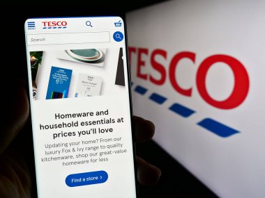 Stuttgart, Almanya - 11-17-2023: İngiliz market perakende şirketi Tesco plc 'nin web sayfası ile akıllı telefon sahibi kişi iş logosu önünde. Telefon ekranının merkezine odaklan.
