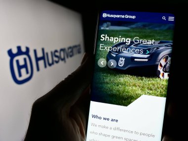 Stuttgart, Germany - 11-22-2023: Person holding cellphone with webpage of Swedish power products company Husqvarna AB in front of business logo. Focus on center of phone display. clipart