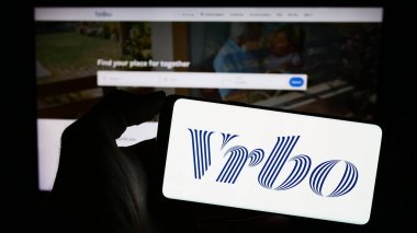Stuttgart, Almanya - 11-29-2023: İnternet sitesi önünde ABD 'li tatil kiralama pazar yeri şirketi Vrbo' nun logosuyla cep telefonu tutan kişi. Telefon ekranına odaklan.