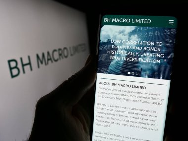 Stuttgart, Almanya - 12-05-2023: Yatırım şirketi BH Macro Limited 'in web sayfası ile birlikte iş logosu önünde cep telefonu tutan kişi. Telefon ekranının merkezine odaklan.