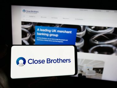Stuttgart, Almanya - 12-06-2023: İngiliz tüccar bankası Close Brothers Group plc 'in logosuna sahip cep telefonu sahibi kişi web sayfasının önünde. Telefon ekranına odaklan.