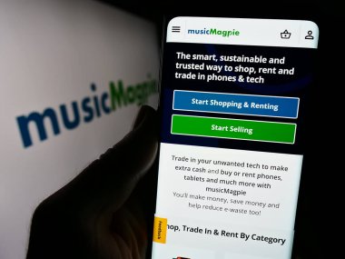 Stuttgart, Almanya - 12-07-2023: İngiliz e-ticaret şirketi MusicMagpie 'nin internet sayfası ile ticaret logosu önünde akıllı telefon tutan kişi. Telefon ekranının merkezine odaklan.