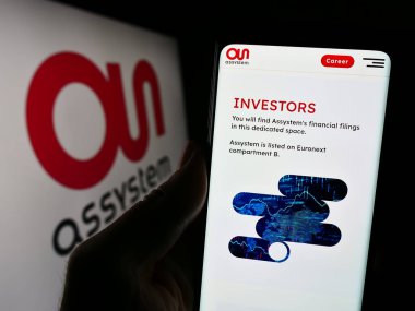 Stuttgart, Almanya - 12-07-2023: Fransız mühendislik şirketi Assystem S.A. 'nın web sayfası ile birlikte iş logosu önünde cep telefonu tutan kişi. Telefon ekranının merkezine odaklan.