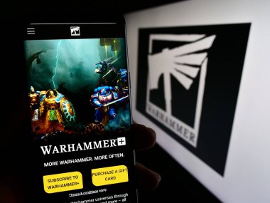 Stuttgart, Germany - 12-12-2023: Person holding smartphone with webpage of video game Warhammer (Games Workshop) in front of business logo. Focus on center of phone display. clipart