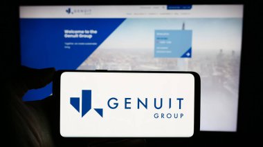 Stuttgart, Germany - 12-12-2023: Person holding cellphone with logo of British piping systems company Genuit Group plc in front of business webpage. Focus on phone display. clipart