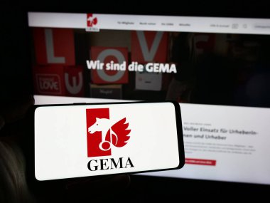 Stuttgart, Germany - 12-14-2023: Person holding smartphone with logo of German collecting society GEMA in front of website. Focus on phone display. clipart