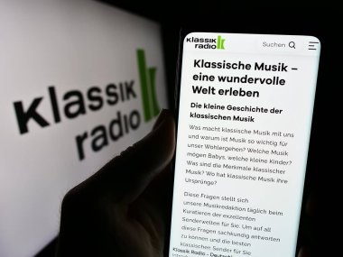 Stuttgart, Almanya - 12-14-2023: Alman yayın şirketi Klassik Radio AG 'nin internet sayfası ile logo önünde cep telefonu tutan kişi. Telefon ekranının merkezine odaklan.