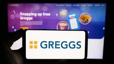 Stuttgart, Almanya - 12-14-2023: İngiliz fırın zinciri Greggs plc 'in logosunun bulunduğu cep telefonu sahibi kişi iş sayfasının önünde. Telefon ekranına odaklan.