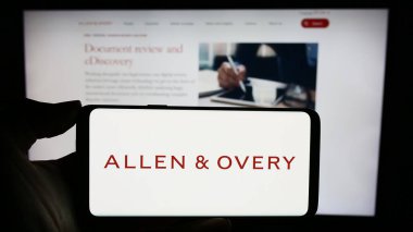 Stuttgart, Germany - 12-17-2023: Person holding cellphone with logo of British law firm Allen and Overy LLP in front of business webpage. Focus on phone display. clipart