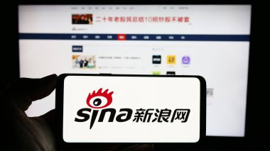 Stuttgart, Almanya - 12-19-2023: Çinli teknoloji şirketi Sina Corporation 'ın logosuyla cep telefonu tutan kişi iş sayfasının önünde. Telefon ekranına odaklan.