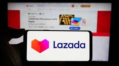 Stuttgart, Almanya - 12-20-2023: Asyalı e-ticaret şirketi Lazada Group 'un logosuyla cep telefonu sahibi kişi iş sayfası önünde. Telefon ekranına odaklan.