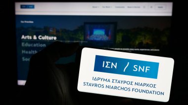 Stuttgart, Almanya - 11-03-2023: İnternet sayfası önünde üzerinde Yunan organı Stavros Niarchos Foundation (SNF) logosu bulunan cep telefonu sahibi kişi. Telefon ekranına odaklan.