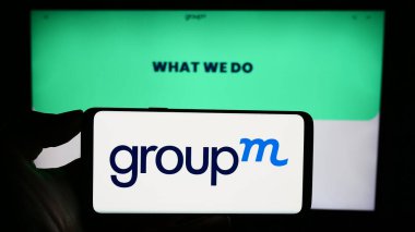 Stuttgart, Almanya - 11-03-2023: ABD 'li medya yatırım şirketi GroupM' nin logosuyla cep telefonu sahibi kişi iş sayfası önünde. Telefon ekranına odaklan.