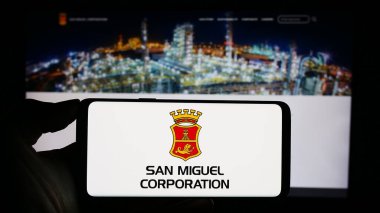 Stuttgart, Germany - 11-08-2023: Person holding cellphone with logo of Filipino company San Miguel Corporation (SMC) in front of business webpage. Focus on phone display. clipart