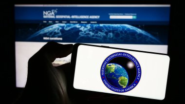 Stuttgart, Germany - 11-11-2023: Person holding mobile phone with seal of US National Geospatial-Intelligence Agency (NGA) in front of web page. Focus on phone display. clipart