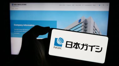 Stuttgart, Almanya - 11-15-2023: Japon seramik şirketi NGK Insulators Ltd. 'nin logosuna sahip cep telefonu sahibi kişi iş sayfasının önünde. Telefon ekranına odaklan.