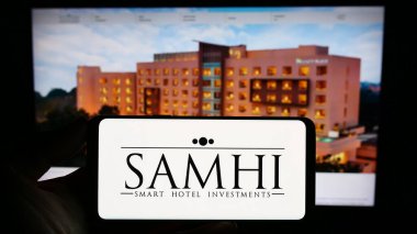 Stuttgart, Germany - 09-28-2023: Person holding smartphone with logo of Indian hotel development company Samhi Hotels Ltd. in front of website. Focus on phone display. clipart