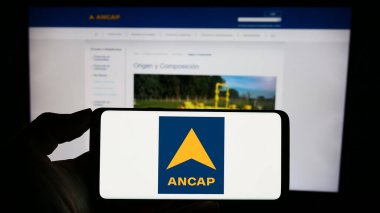 Stuttgart, Germany - 09-28-2023: Person holding cellphone with logo of Uruguayan state-owned company ANCAP in front of business webpage. Focus on phone display. clipart