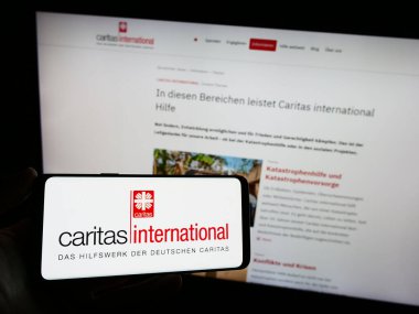 Stuttgart, Germany - 10-08-2023: Person holding mobile phone with logo of aid agency Caritas International in front of web page. Focus on phone display. clipart