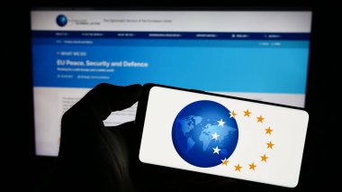 Stuttgart, Germany - 10-16-2023: Person holding mobile phone with logo of EU institution European External Action Service (EEAS) in front of web page. Focus on phone display. clipart