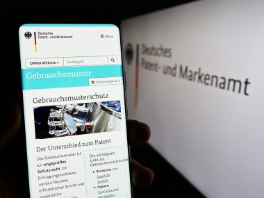 Stuttgart, Almanya - 10-17-2023: Alman Alman ajansı Deutsches Patent-und Markenamt 'ın (DPMA) internet sitesinin logosu önünde cep telefonu tutan kişi. Telefon ekranının merkezine odaklan.