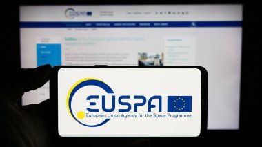 Stuttgart, Germany - 10-17-2023: Person holding mobile phone with logo of European Union Agency for the Space Programme (EUSPA) in front of web page. Focus on phone display. clipart