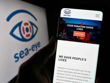 Stuttgart, Germany - 10-27-2023: Person holding cellphone with webpage of German sea rescue association Sea-Eye e.V. in front of logo. Focus on center of phone display. clipart