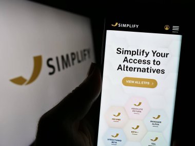 Stuttgart, Almanya - 10-28-2023: ABD 'li yatırım şirketi Basitleştirin Varlık Yönetimi Şirketi' nin web sayfası ile logo önünde cep telefonu tutan kişi. Telefon ekranının merkezine odaklan.