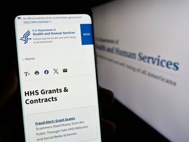 Stuttgart, Almanya - 10-29-2023: ABD Sağlık ve İnsan Hizmetleri (HHS) sitesinin logosu önünde cep telefonu tutan kişi. Telefon ekranının merkezine odaklan.
