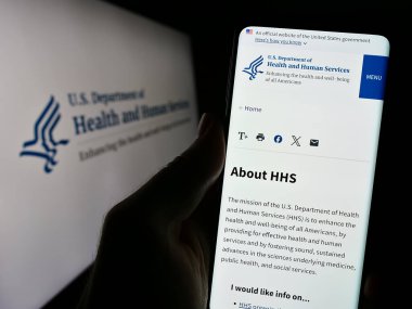Stuttgart, Almanya - 10-29-2023: ABD Sağlık ve İnsan Hizmetleri (HHS) sitesinin logosu önünde cep telefonu tutan kişi. Telefon ekranının merkezine odaklan.