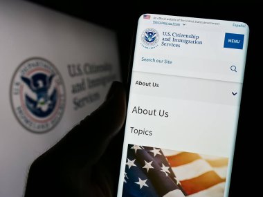 Stuttgart, Germany - 10-29-2023: Person holding smartphone with website of US agency Citizenship and Immigration Services (USCIS) with seal. Focus on center of phone display. clipart