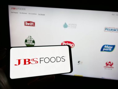 Stuttgart, Germany - 09-10-2023: Person holding cellphone with logo of meat processing company JBS USA Holdings Inc. on screen in front of webpage. Focus on phone display. clipart