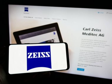 Stuttgart, Germany - 09-17-2023: Person holding mobile phone with logo of medical technology company Carl Zeiss Meditec AG on screen in front of web page. Focus on phone display. clipart