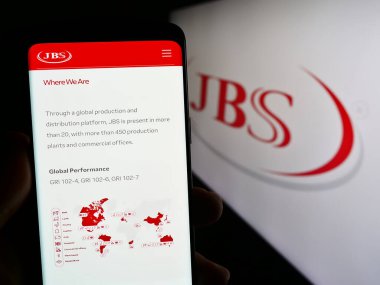 Stuttgart, Germany - 09-17-2023: Person holding smartphone with web page of Brazilian mear processing company JBS S.A. on screen with logo. Focus on center of phone display. clipart