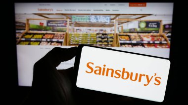 Stuttgart, Almanya - 09-19-2023: İngiliz süpermarket şirketi J Sainsbury plc 'nin logosunu elinde tutan kişi web sitesinin önünde ekranda. Telefon ekranına odaklan.